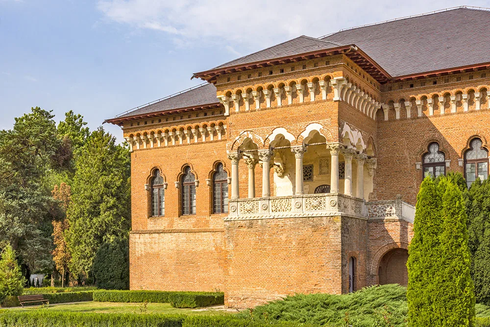 In manicured hedged garden sits a light stone decorative arch building, with arched corbels and trefoil leaded windows and a white columned balcony