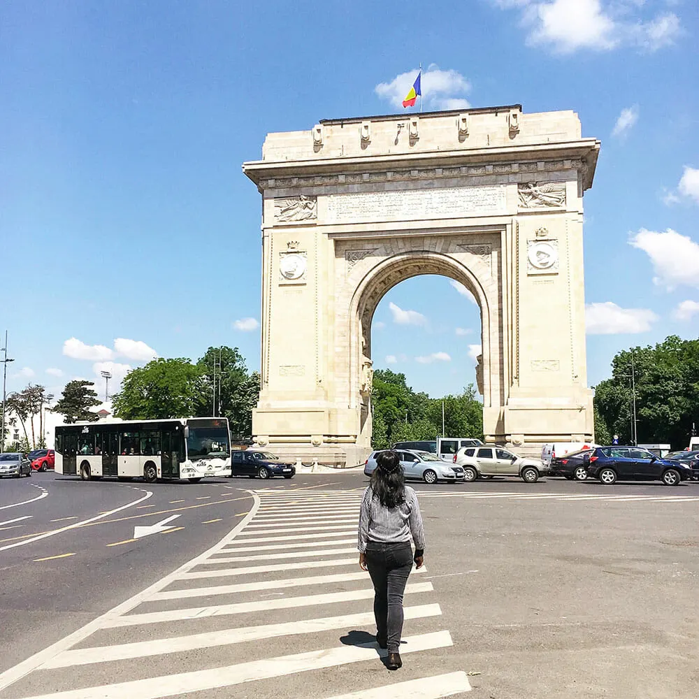 A lady walking towards a tall white stone carved arch, with flag on top, surrounded by cars