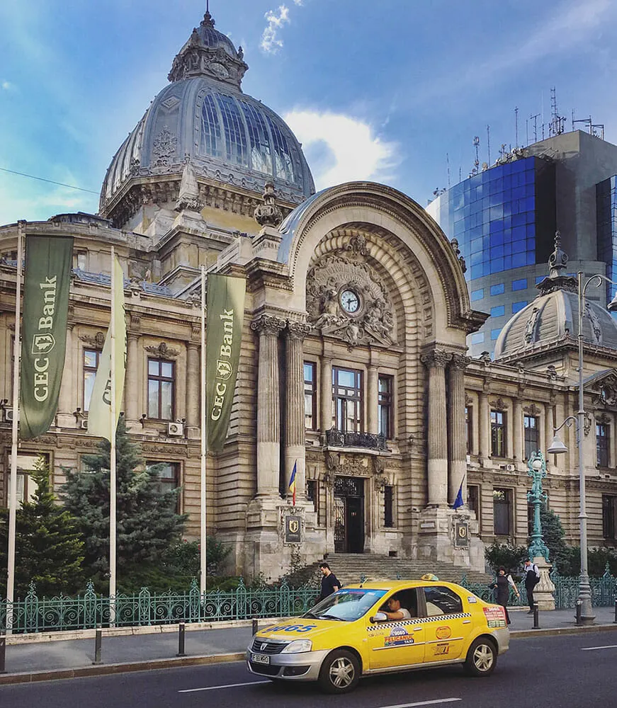 A grand, stone. arch-fronted building with glass atrium top, stands behind flags, green railings and yellow taxi