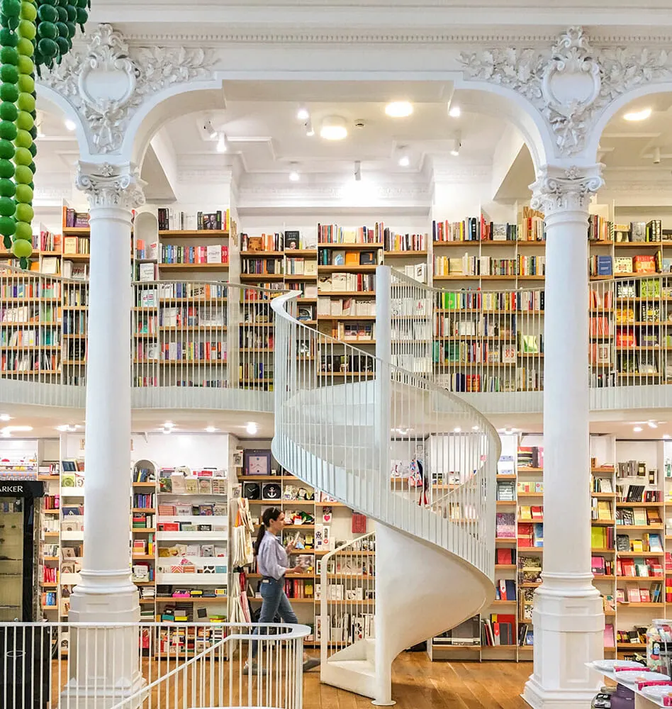 Inside a colourfully stocked, white pillared bookstore, with white railings and spiral staircase