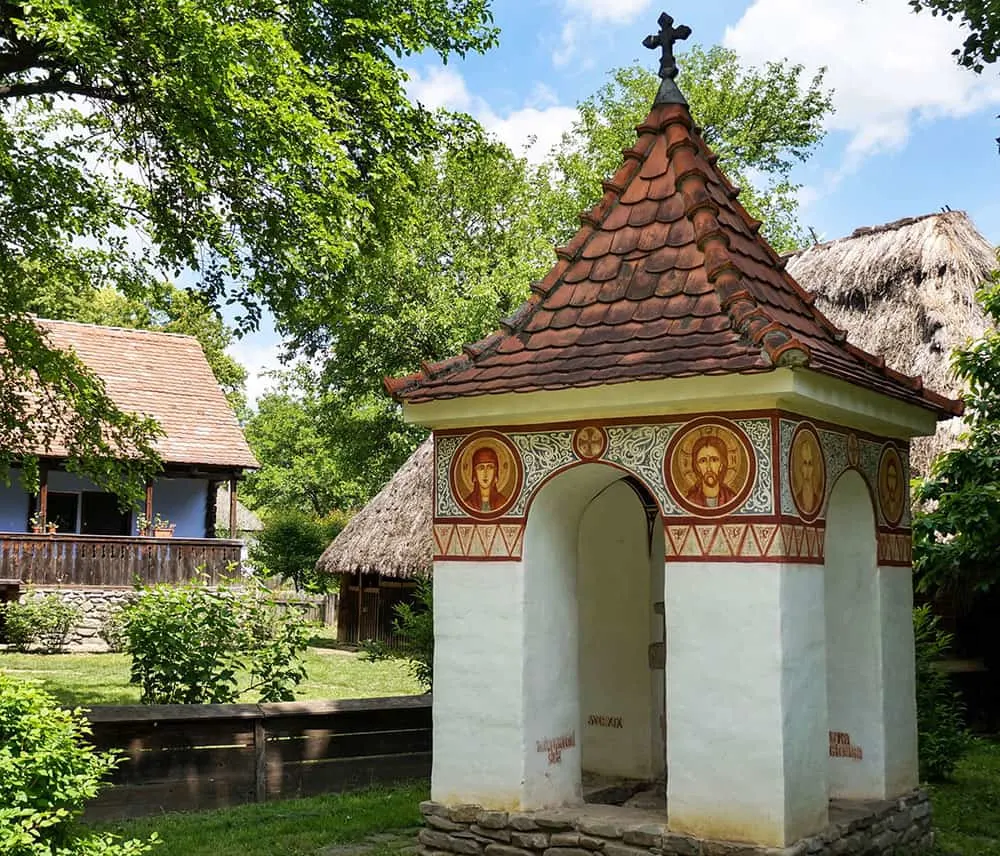 A white shrine with arched sides, frescos and red tiled roof sits in the grounds of a rustic estate