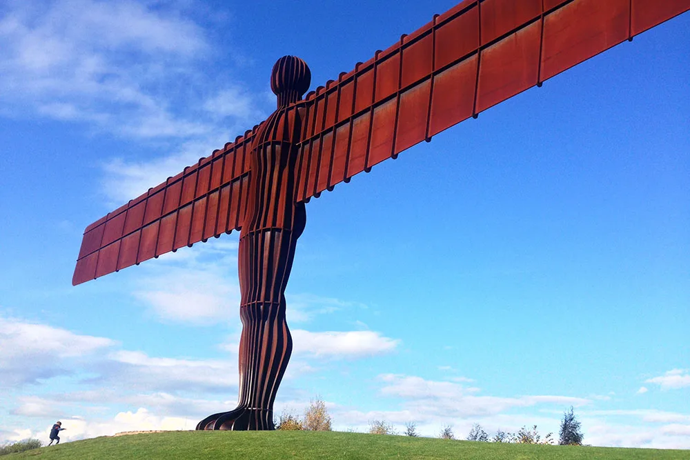 the NewcastleGateshead Angel of the North Newcastle  on top of a green hill featuring its wide wings under bright and clear blue skies
