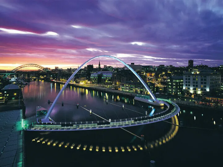 the NewcastleGateshead Millennium Bridge Quayside featuring its amazing curves reflected to the crystal clear river under night and cloudy skies with towns beaming at the side of the river