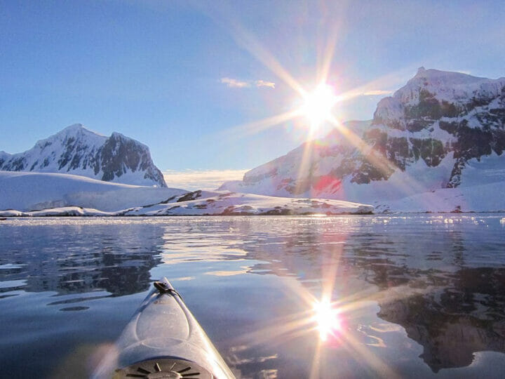 the front of a kayak in icy waters surrounded by snow covered mountains and a bright sunny sky