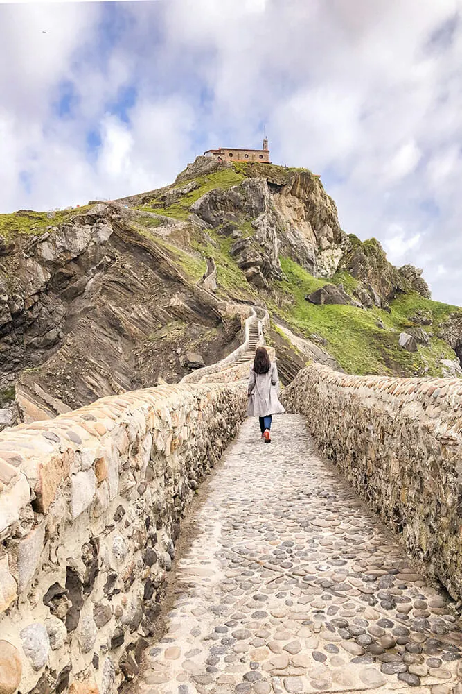 Me walking along a stone-walled path leading across to red buildings on a craggy tied island