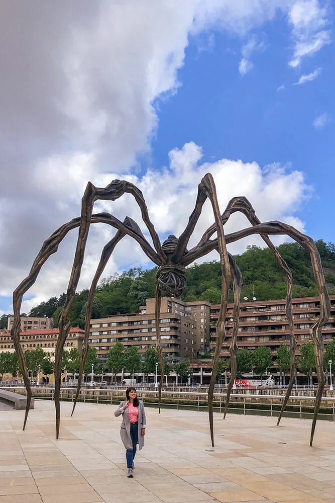A huge, long-legged spider sculpture looms above me on the riverside walkway with red building behind