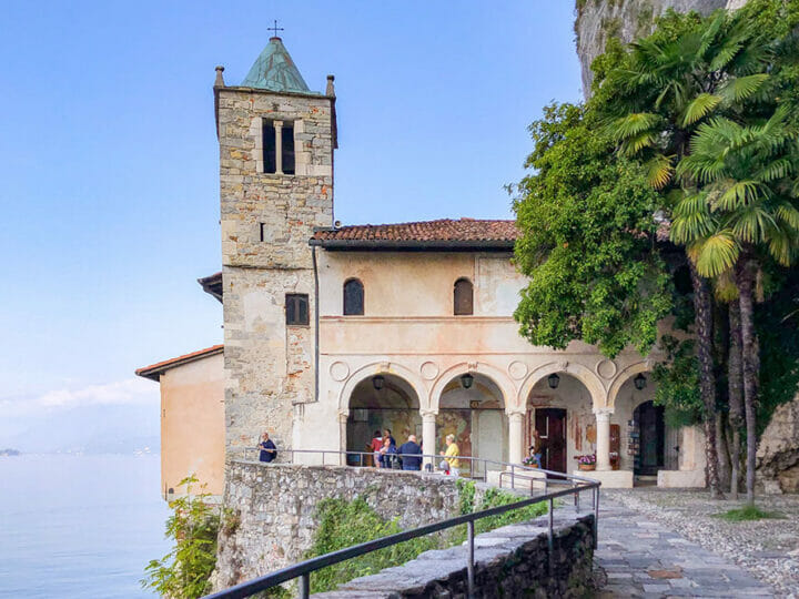 Visitors at a small church with bell tower, situated in the side of a rocky cliff with a lake view.