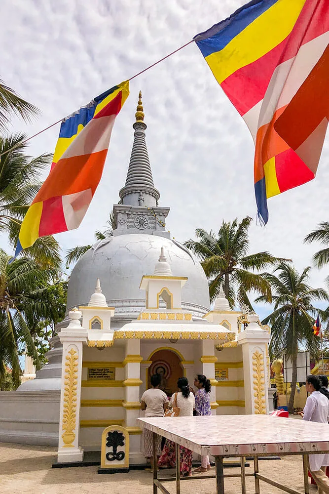 White pagoda with people praying next to it and buddhist flags hanging over it