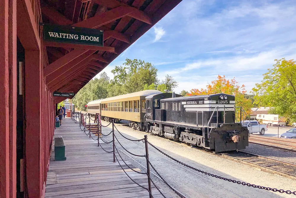 Old train approaching a wooden station platform. There is a sign in the platform reading Waiting Room