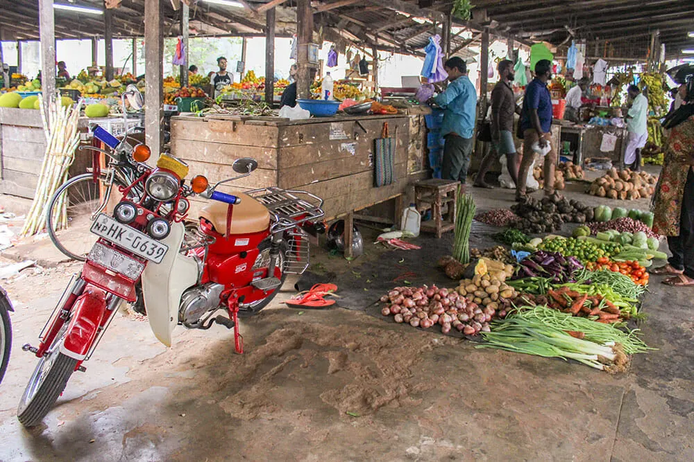 Fresh produce market with wooden stalls and vegetables laid on the floor, with men shopping around and a red motorbike parked next to the stalls
