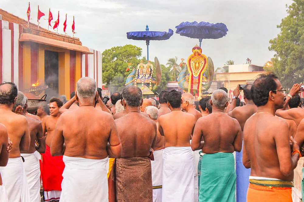 Large group of Hindu men dressed with sarongs and no tops looking away at religious symbols approaching under umbrellas