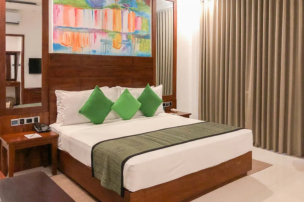 Hotel room with large double bed with white bed linen and three green cushions. There is a colourful painting above the headboard