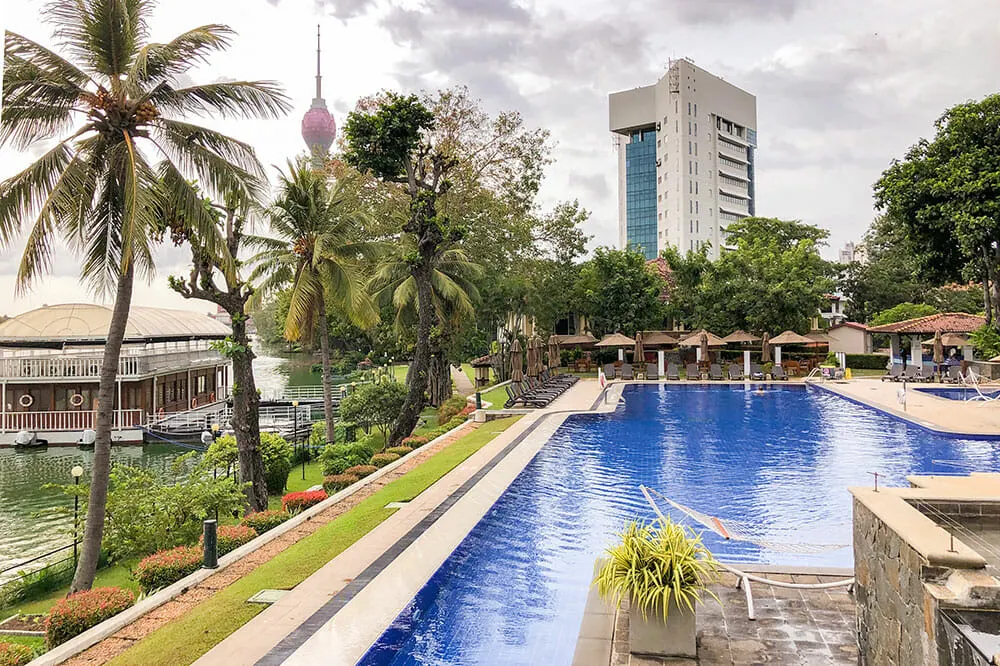 Swimming pool with trees around by a lake with buildings in the background. Cinnamon Lakeside is one of the best eco hotels in Sri Lanka