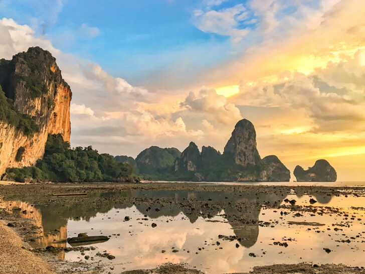 Shallow beach with limestone towers in the distance reflecting on the mirror-like water. The light is golden from the sunset. A beach destination is a must on any Thailand 10 day itinerary