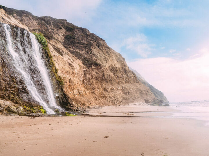 Image of a sandy beach with a waterfall pouring into the beach from a high sandstone cliff