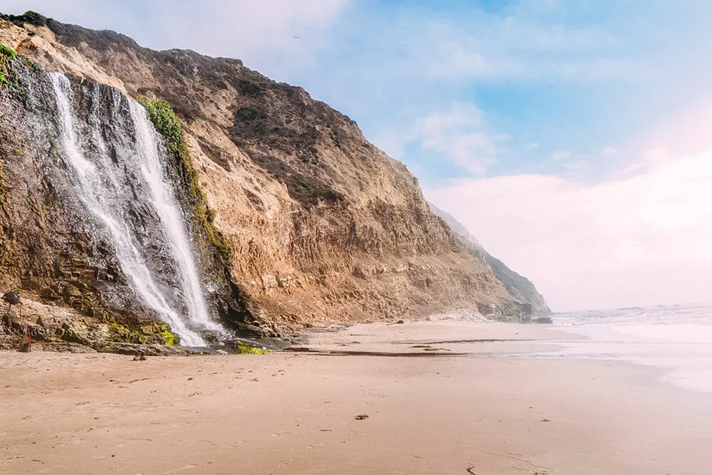 Image of a sandy beach with a waterfall pouring into the beach from a high sandstone cliff