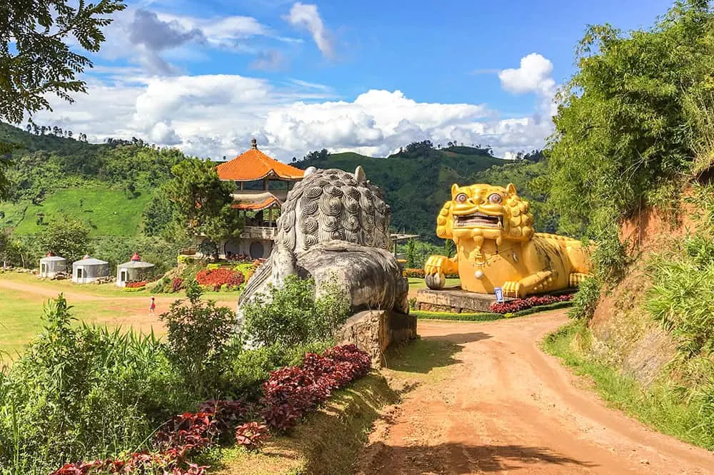 Dirt track leading to a gate garden by two giant Chinese lions, one concrete colour and the other golden. There is a Chinese style building on the other side of the gate and three glamping pods
