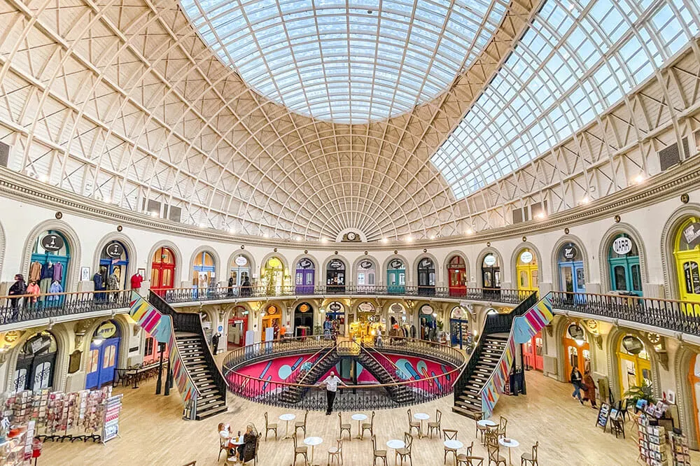 Interior of the Leeds Corn Exchange. There's a cafe with tables and chairs on the ground floor, stair on both side leading up to an upper level lined up with colourful door. The ceiling is white, oval and domed with sections of glass