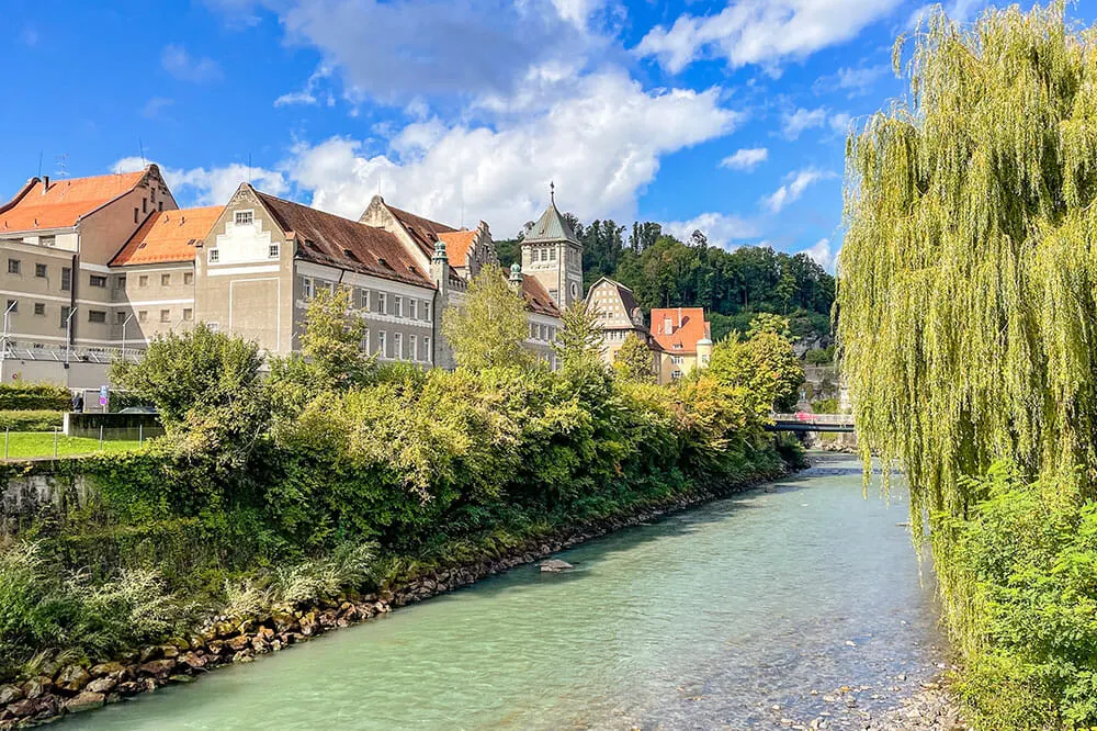 Buildings along the banks of a river with emerald green water. There's a willow tree on the opposite bank