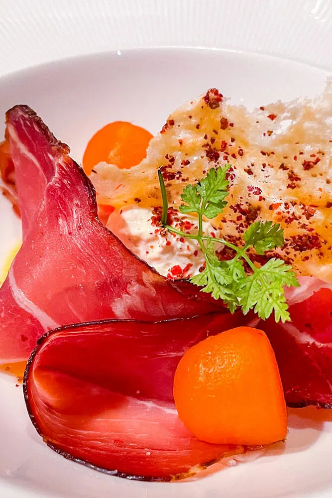 Slices of cured speck with cantaloupe balls and a parmesan crisp on a white plate