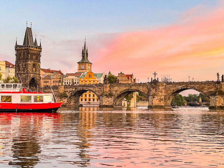 View of a bridge decorated with statues and lights over the Vltava River with people walking and a boat in the foreground