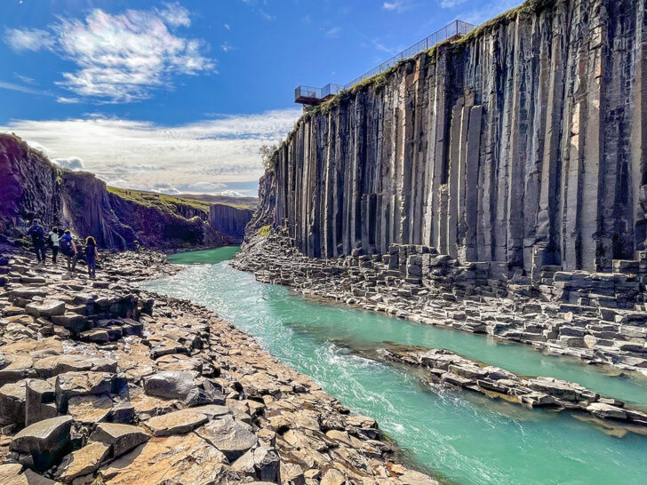 People walking beside clear blue waters with a view of the hexagonal basalt columns lining the canyon walls under bright blue skies and white clouds
