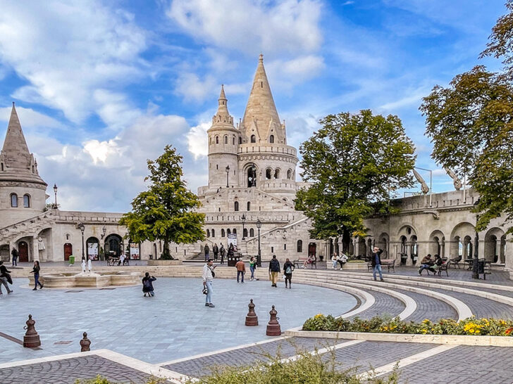 An outdoor space with potten plants and cobbled stoned large stairs with people walking and a view of the a wide building with tower structures of the Fishermans Bastion in the back under bright blue cloudy skies.