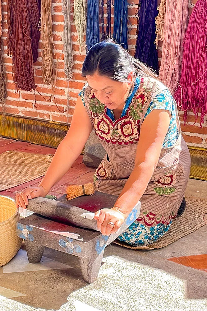 A woman in printed blue and red dress grinding cochinilla on stone with different colors of wool hanged at the bricked wall behind