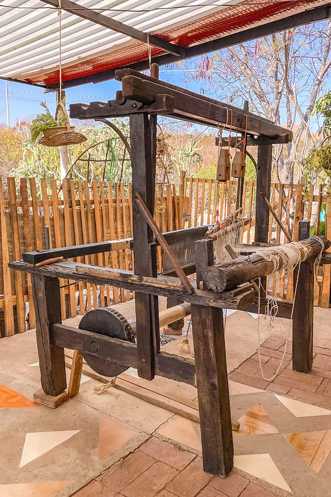 an old wooden loom with white wools on a bricked floor surrounded by wooden fences, tall grasses, and hanging plants