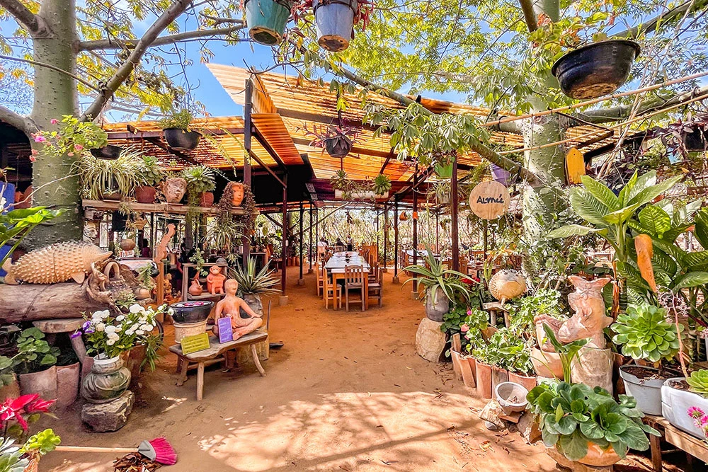 Front of a restaurant with a garden featuring hanging and potted plants and wooden animal sculptures. The entrance leads to the indoor dining area with tables and chairs.