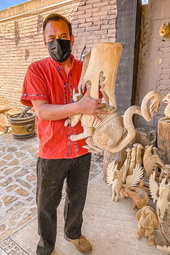 Man in red polo and black pants holding a wooden sculpture surrounded by other animal wooden sculptures on the ground.
