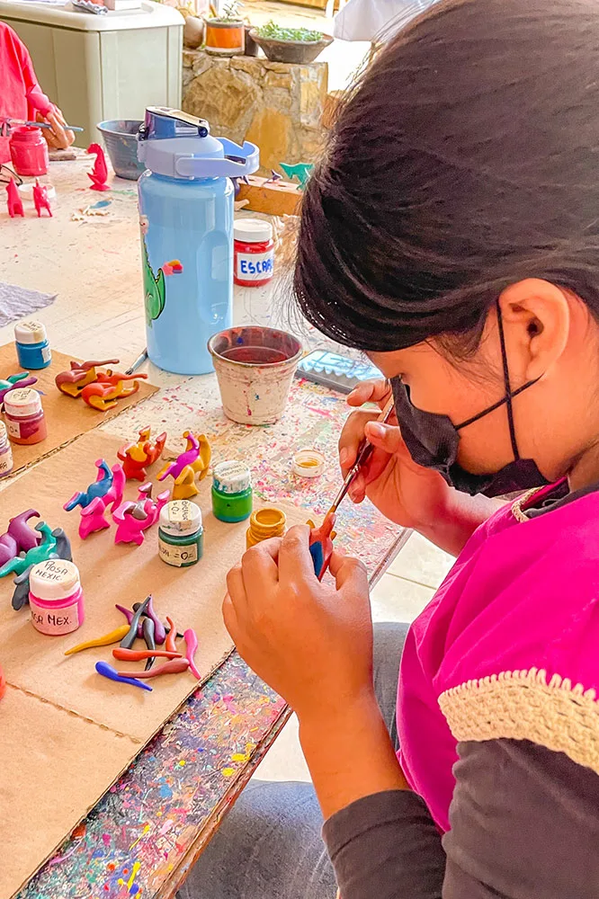 Woman in pink blouse painting small clay figures in red with paints and other clay figures in the background in pink, blue, and green.