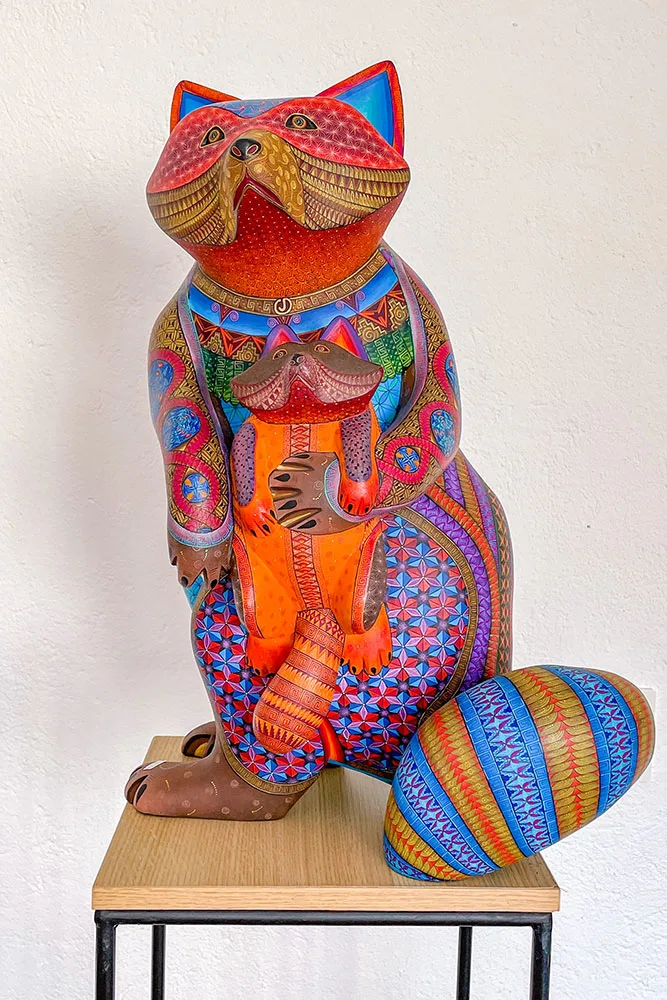 Colorful raccoon sculpture holding a baby raccoon with blue, red, yellow, orange, and other bright colored prints of various shapes and sizes on a wooden square table