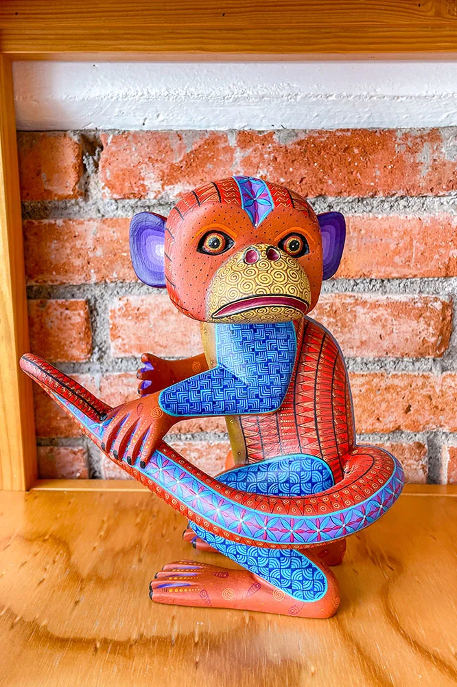 Monkey wooden sculpture holding its tail with blue, red, yellow, orange, and other brightly colored prints of various shapes and sizes on a wooden table, with a brick wall in the background.