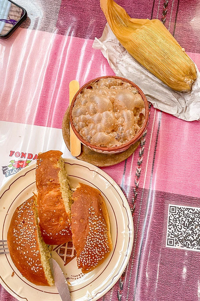 A slice of bread, bubbling cocoa drink, and corn served on a white plate on a pink table.