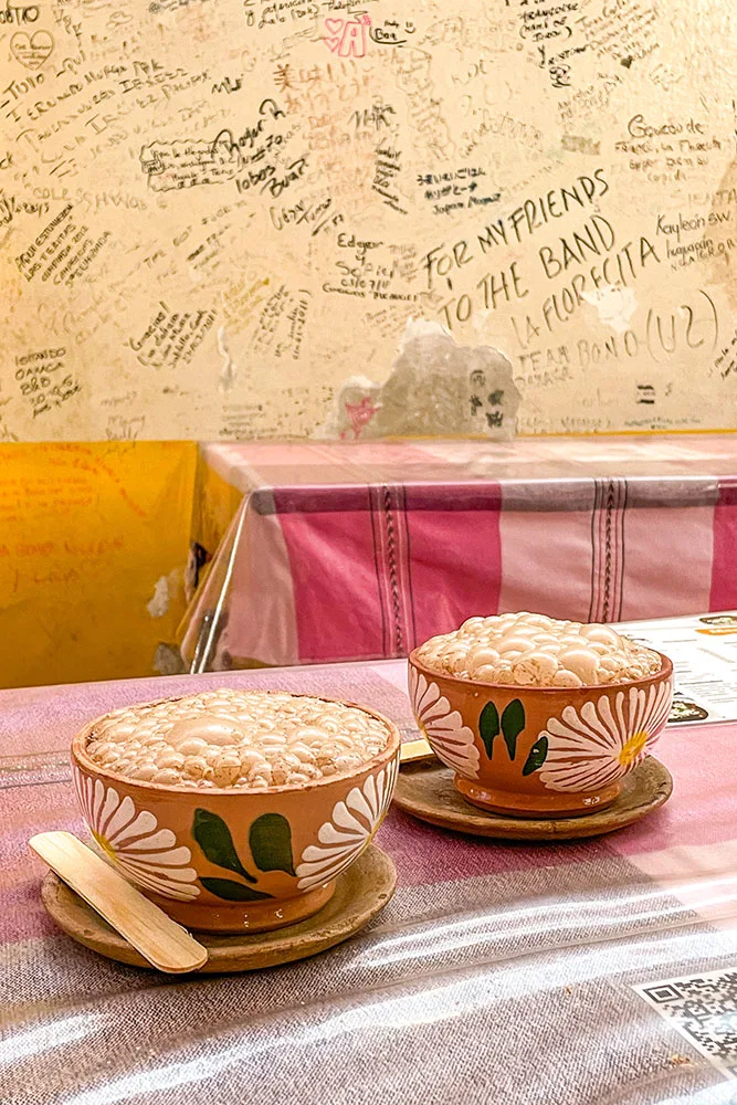 Bubbling Atole drink in brown cups with flower prints on a pink table. Wall in the background filled with texts from customers and visitors.