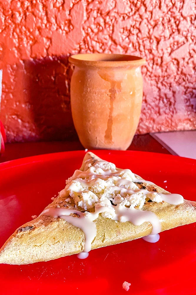 a slice of Tetela with Tepache topped with white sauce served on a red plate and a brown cup on a red table 