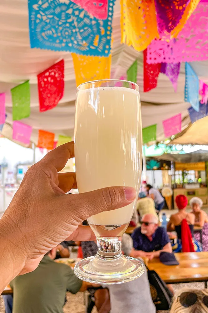 A hand holding a white Pulque drink in a glass with blurry views of people in tables and chairs, and colorful small flags hanging in the background 