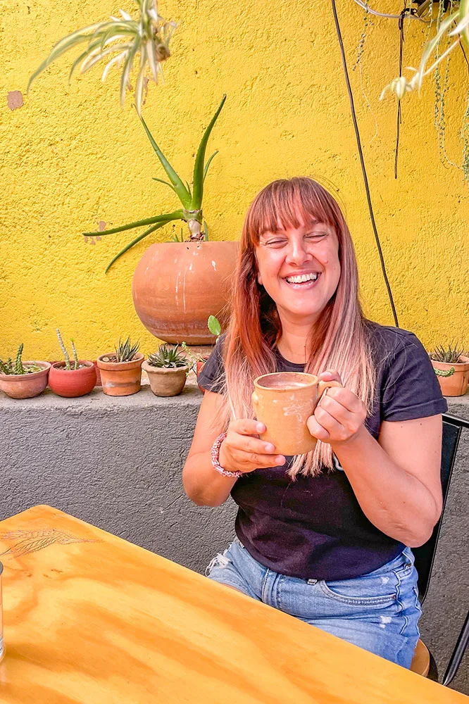 me in black shirt and a pair of jeans holding a brown cup with cacao drink beside a wooden table and potted plants in the background