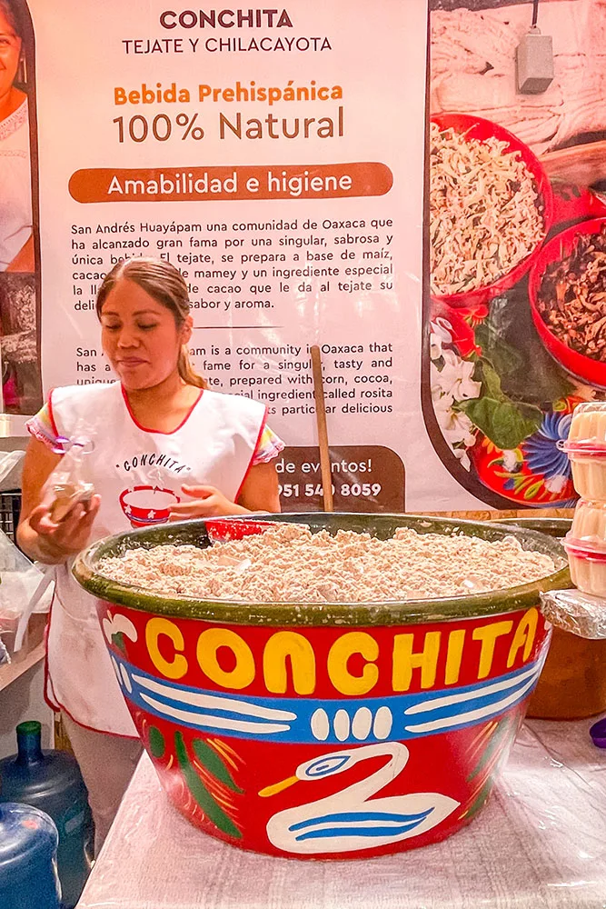 A woman wearing a white apron holding a large cup of Tejate, a traditional Mexican beverage, with text describing the drink in the background. 