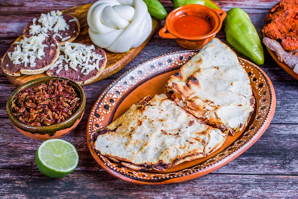 Tlayuda and fried tortilla topped with beans, cream, lime, sauces, and peppers  served on brown plates arranged on a wooden table.