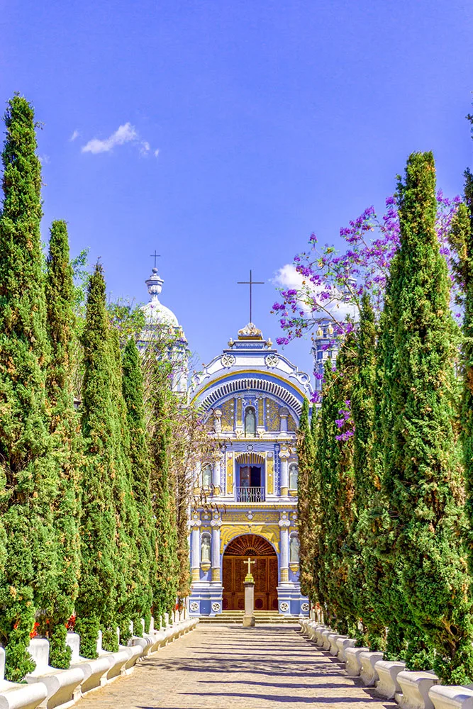 A dirt path with towering trees on both sides leading to Ocotl&aacute;n de Morelos. The blue facade features intricate sculptures and towers, with a prominent cross atop the center of the roof.