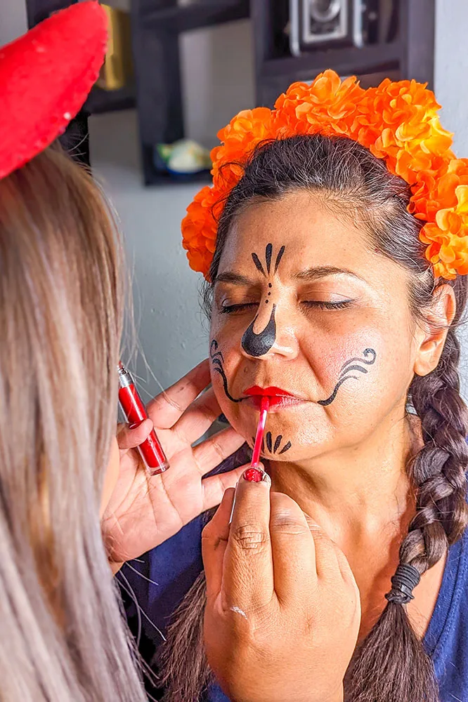 A woman with braids wearing an orange flower headband getting her face painted with black paint by another person