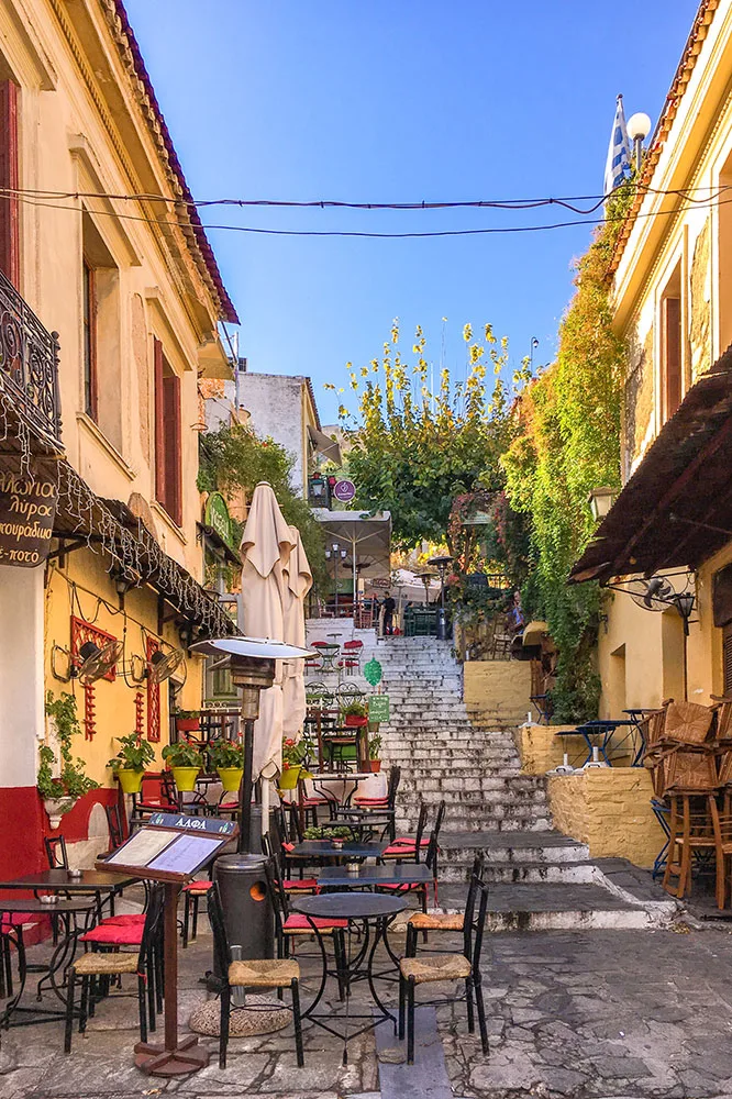A staircase street in between rows of warm colored buildings covered by vines and potted plants under clear blue skies. Black chairs and circle tables with a wooden menu stand in the foreground.