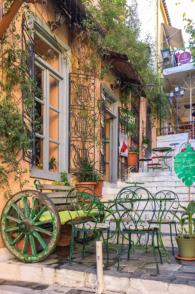A charming staircase street in Plaka adorned with green chairs, tables, and a wooden chair with wheels. The street is lined with orange and white houses featuring large windows covered in vines, complemented by an array of potted plants.