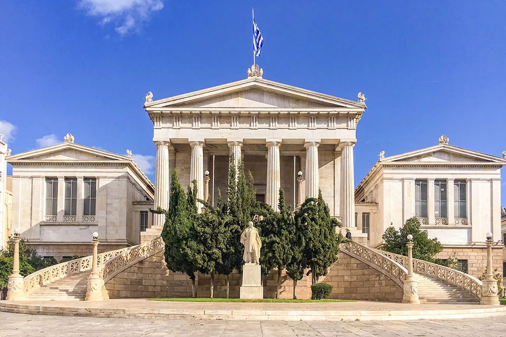 Vallianeio Megaron, a neoclassical masterpiece with grand columns and elegant balconies. Its windows frame views of lush greenery under clear blue skies. Discover this architectural gem among the things to do in Athens.
