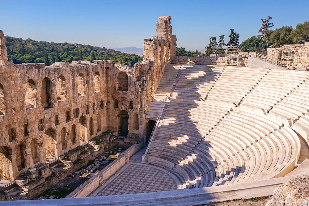 A stunning ancient amphitheater of the Odeon of Herodes Atticus. Its stone seating encircles the stage made of stone bricks with large arched windows under clear blue skies with a view of trees in the back.