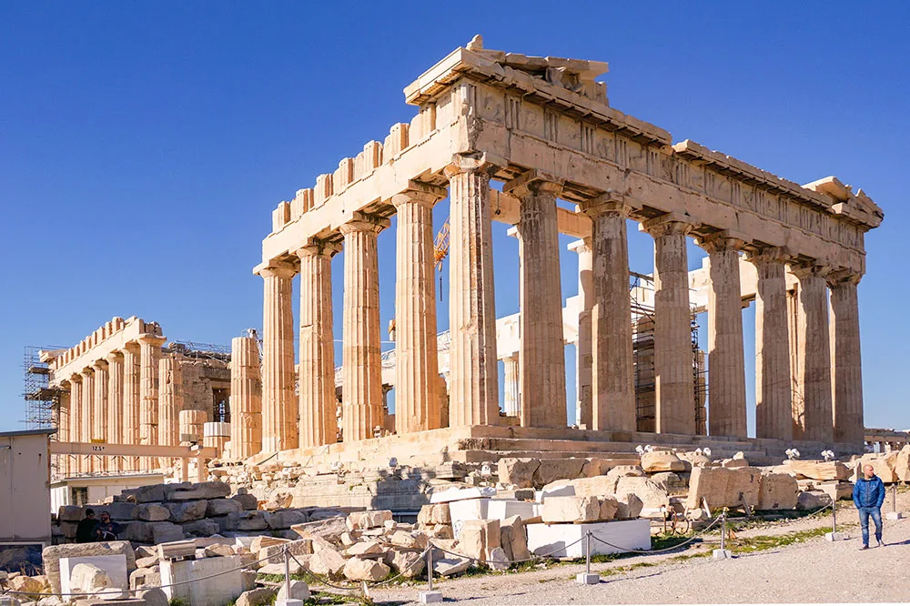 The ruins of the Greek Temple of the Parthenon featuring large Doric columns and intricate marble ruins with a dirt path on the side with a couple of people walking under clear blue skies.