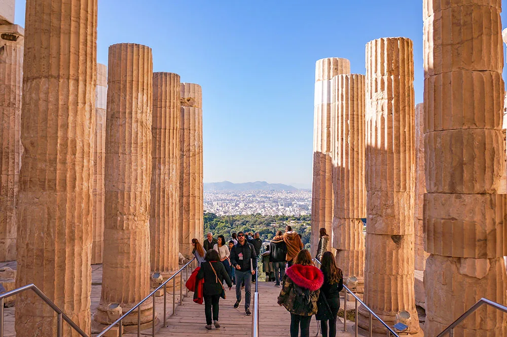 People walking on a wooden path with metal handles surrounded by pillars of the Athens Acropolis under clear blue skies and in the background shows trees and buildings.