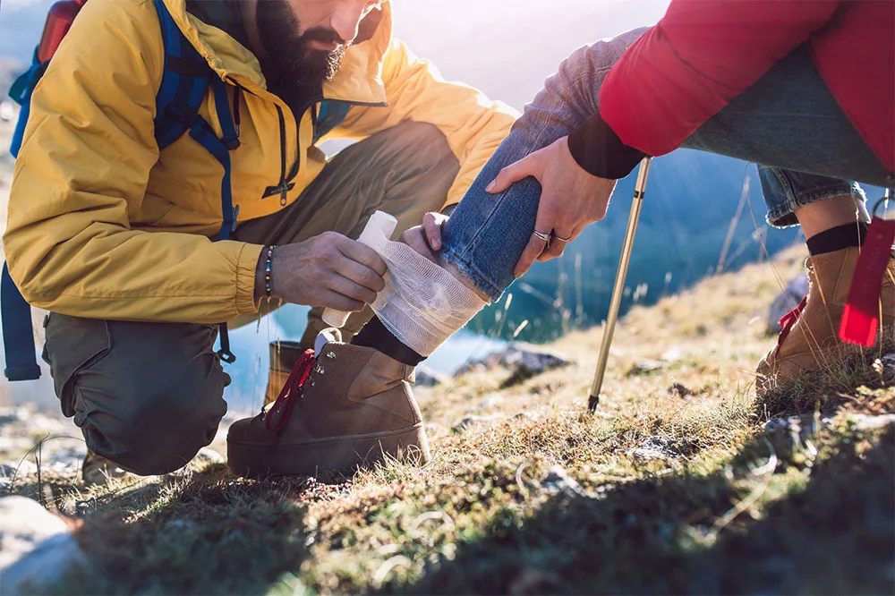 A person in complete hiking gear applying bandage on a leg on another hiker wearing shoes, jeans, and padded red jacket on a grassy field.